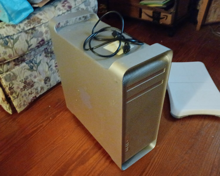 A 2006 Silver Mac Pro computer on a wooden floor. On top of the Mac Pro, a power cable and sticky note are present. An armchair, some shelving, and a Wii Fit Balance Board can be seen in the background.