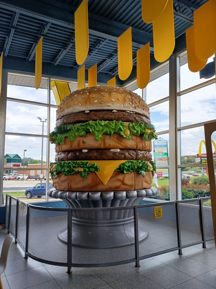 A large statue depicting a Big Mac burger in an inclosed indoor space. The space where the Big Mac statue resides is sectioned off with a fence. A sign on the fence reads Please Do Not Climb Over Fence. Yellow banners can be seen hanging from a black ceiling. A shopping center can be seen through the glass in the background, as well as a sign advertising the McDonalds Big Mac Museum.