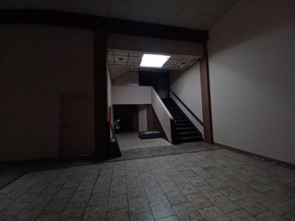 A liminal space that is dark, but contains a staircase with a single light fixture illuminating a limited portion of the space. The floor is made of tile, and the ceiling appears to be made out of panels with dark borders. At the top of the stairs, there appears to be an area that's sectioned off with glass. At the bottom of the stairs, there appears to be a dark hallway leading somewhere. There's also a shallow, blue tub that appears to contain an unknown white object or assortment of objects. A fire extinguisher can be seen mounted to a wall, with a piece of wood and gold-foil ballon leaning up against it. To the left of the dark hallway, and to the left of a brick column, there's a door with a tiny bit of light appearing from underneath.