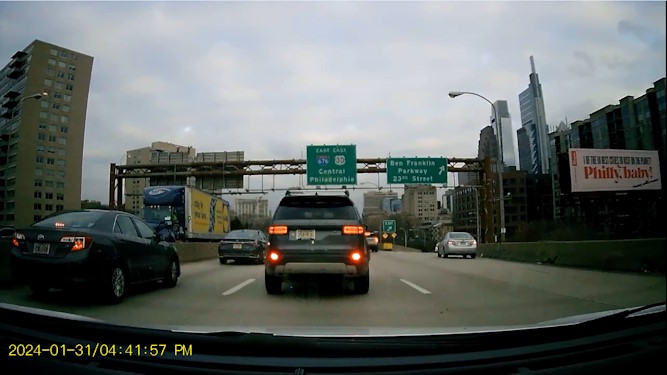 A screenshot of dashcam footage of the Center City Expressway in Philadelphia. Several tall buildings can be seen in the background. A billboard to the right shows text on a white background, with the phrase Philly Baby written in large red print. There are two highway exit signs in the center of the screenshot. One sign is for East Interstate 676 US Route 30 East Central Philadelphia. The second exit sign is for the Ben Franklin Parkway and twenty third Street. Several cars are seen in front of the camera's view. The date marker January thirty first 2024 slash four fourty one fifty seven PM is shown in the bottom-left.