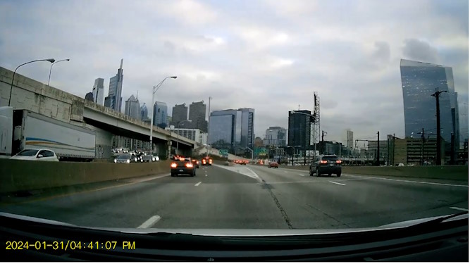A screenshot of dashcam footage of the Philadelphia skyline taken from the perspective of a car on a highway ramp. Several skyscrapers are shown prominently in the center and right of the photo. Several cars can also be seen on the highway. The date marker January thirty first 2024 slash four fourty one oh seven PM is shown in the bottom-left.