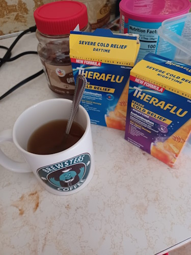 A mug with the Brewster's Coffee logo from Animal Crossing on a countertop. The mug appears to be filled with Theraflu, a hot liquid medicine used for treating cold and flu symptoms. To the right of the mug are two boxes of Theraflu. One for daytime use, and one for night time use. A container of instant coffee and a container of drink mix can be seen in the background.